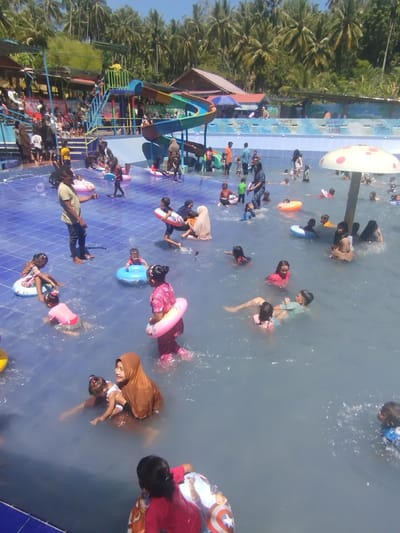 The image shows a crowded outdoor water park in Palohseulimeng, Indonesia. Many children and adults are in the pool, swimming or wading. Some are using inflatable rings. A colorful water slide is a prominent feature of the scene. Behind the pool, there are palm trees and buildings. The sky is clear and blue.