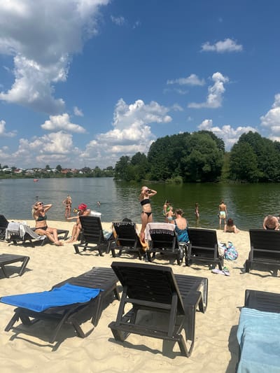 The image depicts a sunny day at a sandy beach next to a body of water, possibly a lake or river, with lush green trees and a village visible in the background. The sky is a vibrant blue with fluffy white clouds. Several people are present, some sunbathing on black lounge chairs scattered on the sand, while others are in the water.
In the foreground, a woman in a dark bikini is lying on a lounge chair, facing the sun, with her hands resting on her head. Another woman in a green bikini is also sunbathing nearby. To the left, a woman in a black bikini stands with her hands on her hips, looking towards the water. Further back, several individuals are visible in the water, some appearing to swim or wade. A man in patterned swim trunks is standing at the edge of the water.
Various objects are present on the sand, including beach towels of different colors, a patterned beach bag, and flip-flops. The black lounge chairs are a prominent feature of the scene, suggesting a recreational area. The overall atmosphere appears relaxed and leisurely, consistent with a warm summer day at a beach. The presence of the village and dense trees indicates a natural, possibly semi-rural setting. The time of day is likely midday or early afternoon, given the bright sunlight and shadows. No visible text is present in the image.