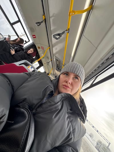 The scene depicts the interior of a public bus in Плишки, Russia, on an overcast winter day. In the foreground, a female subject is captured from a low angle, looking upwards and to the right with slightly parted lips. She wears a grey ribbed knit beanie and a dark grey puffer jacket; a black textured handbag rests on her left side. Behind her, on the left side of the bus, two other female passengers are seated. One is observing a smartphone, while the other is looking forward. The bus interior features red fabric seats, white ceiling panels, and prominent yellow metal grab bars with black handgrips along the ceiling and side. A rectangular light fixture is visible on the ceiling, and a red emergency symbol is on the upper wall. Through the windows, a blurry, snow-covered landscape with bare trees and a partially visible white vehicle suggests outdoor winter conditions.