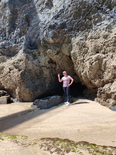 A full-shot image shows a person standing on a sandy beach in front of a cave formed by large, rough rock formations. The person, wearing a striped t-shirt, dark pants, white sneakers, and a white baseball cap, stands with their left hand on their hip and their right hand extended, forming a peace sign. The cave entrance is dark, contrasting with the brightly lit beach. The sandy beach is wet in some areas, with patches of seaweed visible. Shadows from the rock formations cast a diagonal line across the sand. The rocky landscape is rugged and textured, with some areas appearing darker and smoother than others. In the background, the cave extends into darkness. The location is the beach in Newquay, United Kingdom.