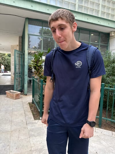 A young male, appearing to be a teenager or young adult, stands on a light-colored tiled pathway in front of a building entrance at an ORT Learning Center in Ness Ziona, Israel. He has short, light brown hair and fair skin. He is wearing a dark blue short-sleeved athletic t-shirt with a white circular logo on the left chest that reads "ORT Learning Center Ness Ziona" in Hebrew. A dark blue backpack is worn over his shoulders, and a black digital watch is on his left wrist. He wears dark blue shorts or athletic pants, with both hands fisted at his sides. His head is tilted slightly down, eyes looking downwards and to his left, with his lips slightly pursed.

The background features a building with a partially open light green metal door leading into an interior. To the left of the door, a rectangular terra cotta planter is on the ground. The building's facade above the entrance includes light green window frames and sections of glass blocks. To the right of the subject, a green metal fence runs alongside a small patch of dirt with sparse green vegetation. In the deeper background, to the far left, another individual wearing a light-colored shirt and dark pants is visible walking away from the camera. Lush green trees and foliage are present in the distance. The scene is illuminated by natural daylight.