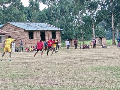 A soccer game unfolds on a dry, grassy field in a rural setting. Players in red and yellow jerseys compete, while spectators watch from the sidelines. A brick building with a tin roof serves as a backdrop, alongside lush trees and a small shed. The atmosphere suggests a local or amateur game, with a community gathering to enjoy the sport.