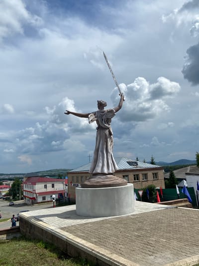 A full-body, low-angle shot captures a statue of a woman holding a sword aloft against a cloudy sky. The statue is positioned in the foreground, centered within a paved plaza. The woman is depicted in a flowing robe, with her right arm raised holding a long, slender sword, and her left arm outstretched. Behind the statue, a multi-story brick building with a sloped roof is visible, along with other buildings and a distant hillside. The sky is filled with dramatic, dark clouds, suggesting an overcast day. To the left of the statue, a street with a car and a lone pedestrian is visible. Several flags, including red, white, and blue, are planted in the ground to the right of the statue. The foreground is a paved area with interlocking concrete blocks, bordered by a stone wall and grass. The overall scene is static, with no discernible action or interaction between subjects, beyond the presence of the pedestrian. The location is identified as the city of Askarovo, Russia.