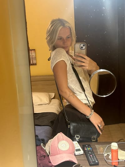 A young woman with blonde hair is taking a mirror selfie in what appears to be a hotel room. She is wearing a light-colored, textured top and has a black crossbody bag slung over her shoulder. Her left hand is holding her phone, which is in a light-colored case, and her right hand is resting on her bag. She is wearing bracelets on her left wrist and a ring on her right index finger.

In the reflection, we can see a small circular mirror hanging on a dark wall above her. On a surface to the right, there are several objects including a remote control, a white remote-like device, a pink baseball cap, and an orange and white cylindrical object. Behind her, in the reflection, are what appear to be hotel room furniture, including a bed with white linens and wooden closet doors. The room has a warm, yellowish tone from the lighting.

The overall mood seems casual and relaxed, perhaps a travel photo taken during the day. The image is oriented vertically, but the contents are viewed in a mirror reflection, so they appear sideways. There is no visible text in the image.