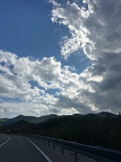The image captures a majestic mountainous landscape, likely in Očinići, Montenegro, under a dramatic sky. The scene is dominated by a vast expanse of dynamic clouds and a towering, forested mountain range.
The sky, occupying the left and upper portions of the frame, is filled with a mix of bright white cumulus and darker stratocumulus clouds. Patches of clear, deep blue sky are visible between the cloud formations, particularly in the lower left. The sun, though not directly visible, brightly illuminates the edges and undersides of many clouds, creating a luminous effect and suggesting a bright, partially cloudy day.
On the right side, a steep, densely vegetated mountain rises sharply, covered in dark green foliage, indicative of a rugged, natural environment. Its peak extends beyond the top of the frame. In the foreground, a paved road curves from the bottom right towards the middle right, alongside the mountain. A robust metal guardrail runs parallel to the road, providing a sense of scale and safety for the winding path. White road markings are discernible on the dark asphalt surface.
The time of day appears to be daylight, with strong natural illumination. No people, vehicles, or animals are visible, emphasizing the grandeur and solitude of the natural environment. A small, white rectangular sign is present near the roadside at the base of the mountain, but its text is indecipherable. The overall impression is one of serene yet imposing natural beauty.