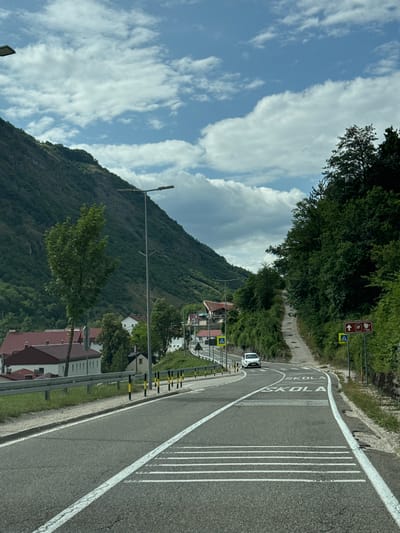 This is a photograph of a road leading into a town nestled in a valley in Šavnik, Montenegro. The road is paved and has white lane markings. There are speed bumps painted on the road with the word "SKOLA" (school) written on them. A white car is driving away from the camera. On the left side of the road, there are buildings with red roofs, a metal guardrail, and trees. On the right side of the road, there is a steep, tree-covered hillside and road signs indicating a school and possibly a parking area. The sky is partly cloudy. The scene appears to be taken from a vehicle moving along the road.