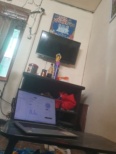 The image shows a room in a house located in Kecamatan Jombang, Indonesia, at coordinates -7.519 latitude and 112.236 longitude. The room is sparsely furnished and contains a window on the left side of the image. The window is covered by a white curtain with grey and black stripes. There is an open window in the back corner, a flat screen TV mounted on the wall, and a shelf with various items on it, including a trophy, bottles, and a can. There is a black and red bag on the shelf.  The room also contains a table with a laptop open on it. The laptop screen displays a graph. There is a glass top table in the foreground and a blue patterned blanket on the floor in the foreground. The ceiling is white. The walls are painted white. There are no people visible in the room. The scene is lit by natural light coming in through the window. The room appears to be a living room or a study.