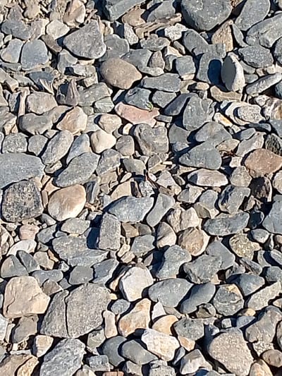 The image shows a close-up, overhead view of a pile of grey and tan rocks and gravel. The rocks are of various sizes and shapes, with some being smooth and rounded, while others are jagged and angular. The lighting suggests that the sun is high in the sky, casting shadows on the rocks. There are no people or any signs of human activity visible in the image. This scene could be found in any location with loose gravel, including the city of Magadan, Russia, which is known for its rugged terrain and gravel roads.