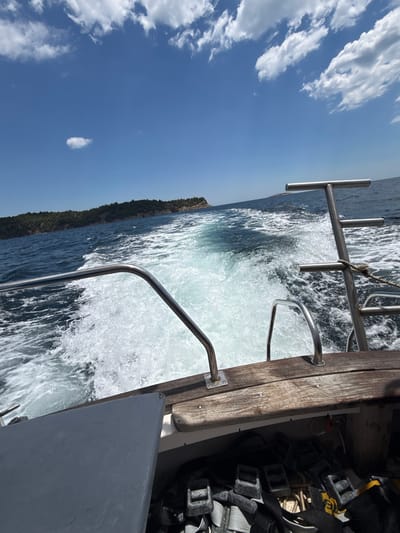 The image captures a view from the stern of a boat moving through the sea, creating a white wake. In the distance, a lush green, tree-covered island coastline is visible under a bright blue sky dotted with white, fluffy clouds. The boat itself appears to be made of wood and metal, with a railing and a gray surface in the foreground. Below the railing, some equipment, including straps and ropes, is stored. The scene depicts a sunny day at sea, likely near the coast of Potos, Greece, given the context of the location. There are no people visible in the image.