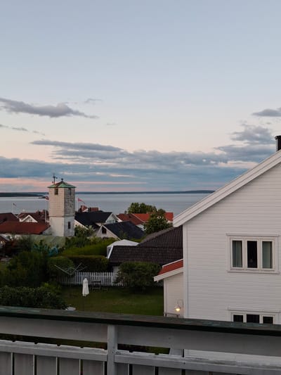 The image depicts a view overlooking Åsgårdstrand, Norway, at dusk. In the foreground, a white wooden railing with weathered green trim is visible. Beyond the railing, a residential area with several houses is spread out.
The houses feature a variety of roof colors, including red and dark gray. One prominent structure in the middle ground is a white, square tower with a dark green roof, resembling a lighthouse or a small observation tower. Two Norwegian flags are flying from poles near this tower.
The houses are surrounded by lush green trees and bushes. A neatly maintained lawn with a white picket fence is visible in the middle ground, with a clothesline and a closed white umbrella. In the foreground to the right, a section of a white, wooden house with horizontal siding and a visible window is present. A single outdoor lamp with a warm glow is also visible on this house.
In the background, a body of water, likely the Oslofjord, stretches towards the horizon. The sky above is a soft blend of pale blue and light pink, with scattered gray clouds, indicating the late evening light. The overall scene conveys a peaceful, serene coastal atmosphere.