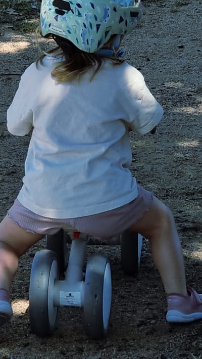 A person, seen from behind and from the waist down, is sitting on a balance bike. They are wearing a light-colored t-shirt, pink shorts, and pink shoes. The balance bike has two wheels in the back and one in the front. The background is a granular, dirt-like surface. The image appears to have been taken outdoors.