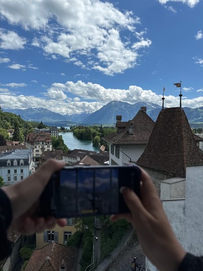 A person's hands hold a smartphone horizontally to take a photo of a panoramic view of Thun, Switzerland. The city is nestled beside a wide, turquoise river that flows between lush green banks and leads to a distant lake. Beyond the city and river, majestic, snow-capped mountains rise against a bright blue sky dotted with fluffy white clouds. The foreground features the tiled rooftops of historic buildings, including a prominent castle tower with decorative flags. The phone screen reflects the scene being captured, showing a portion of the sky and mountains. The overall atmosphere is one of natural beauty and historical charm.