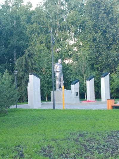This media file shows a monument in a park setting. The central subject is a silver statue of a man in uniform, possibly a soldier or a historical figure, standing on a pedestal. Behind the statue are three tall, slender, light-colored structures with dark tops, likely memorial plaques or panels. To the left of the statue, there's a smaller evergreen tree and a lamppost. The background is dominated by lush green trees, suggesting a daytime scene with moderate foliage. The ground in the foreground is covered in green grass, with some patches of exposed soil and smaller plants.

The overall scene appears to be a war memorial or a commemorative site, likely in a public park. The presence of the statue and the memorial panels points to a tribute or remembrance. There are no people visible in the frame, and the setting is peaceful and solemn. The lighting suggests it's daytime, likely morning or late afternoon, given the soft illumination. The weather appears to be pleasant, with clear skies implied by the brightness and the abundant greenery.

Notable details include the metallic sheen of the statue, the angular design of the memorial panels, and the lamppost providing illumination. A red star-shaped object, possibly a wreath or decoration, is visible near the base of the memorial panels. To the far right, a part of an orange bench and a trash bin are visible, indicating the area is maintained and accessible to the public. No visible text is discernible in the image. The location context provided is Mikhailovka, Russia, which places this monument within a specific geographical and cultural setting.