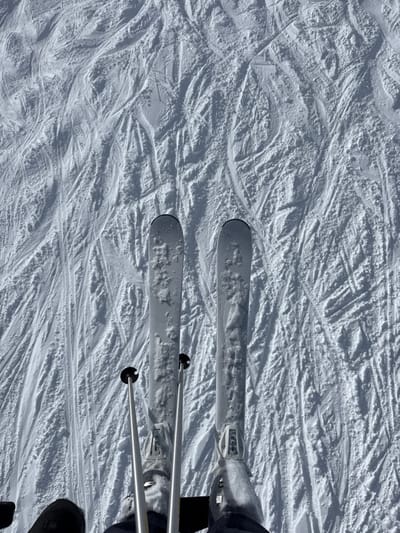 The image depicts a first-person, downward perspective of a skier positioned on a snow-covered slope in Saint-Martin-de-Belleville, France. The foreground features the tips of two white alpine skis, partially covered with crystalline snow, with their respective bindings and the top portions of black ski boots visible. Two metallic grey ski poles with black baskets are positioned between the skis, resting on the snow. The expansive background consists entirely of a white, undulating snow surface heavily marked by numerous crisscrossing ski tracks, indicating a frequently used piste. The scene is illuminated by natural light, casting subtle shadows that highlight the snow's texture. No other individuals are present in the visible frame.
