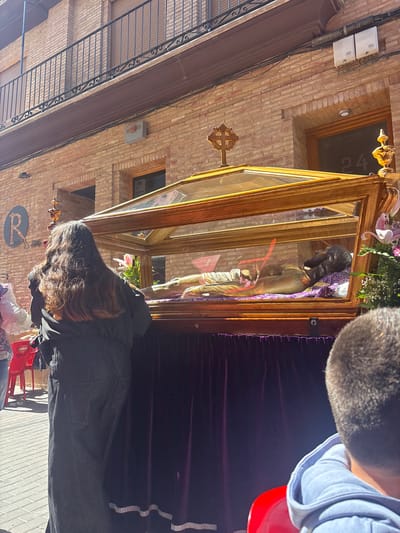 The image shows a religious procession in the town of Mallén, Spain. A wooden and glass case containing a statue of Christ is being carried through the street. A person in a black jacket with long dark hair stands near the case. A partial view of another person's head and shoulder is visible at the top right of the frame. The architecture of the town is visible in the background, including brick buildings with ornate windows. There is a sign with the letter R on a building. The street is paved with gray blocks.