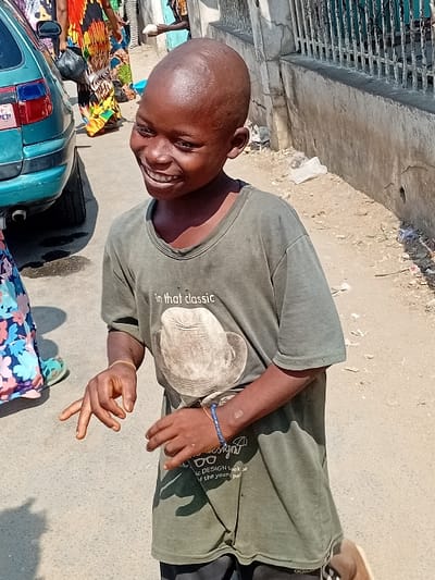 A smiling young person with a shaved head stands outdoors, wearing a green t-shirt featuring a hat graphic. The individual makes a hand gesture. A teal car is visible in the foreground. Behind the person, what looks like a building or gate is visible on the left, as well as some litter. The surrounding area looks like a street.