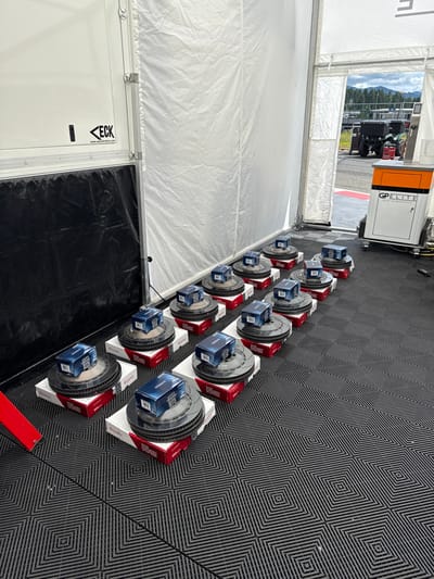 A row of brake rotors on white stands with red accents are displayed inside a tent on a black and gray geometric floor. Blue boxes sit atop each rotor. Behind the rotors, a doorway reveals a race track with mountains in the distance. A GP ELITE branded generator sits near the doorway. To the left, a white wall with an ECK logo and black sheeting is visible.