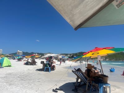 The image displays a vibrant and bustling beach scene on a clear, sunny day. The perspective is taken from under a light-colored beach umbrella, a corner of which is visible in the upper right portion of the frame.
In the foreground, slightly to the right, two beach chairs are set up. One chair is occupied by a person seen from behind, wearing a straw hat and appearing to relax under the shade of a brightly colored umbrella. This umbrella features distinct sections of red, yellow, and green fabric, with the number "6" clearly visible in black paint on its yellow section. Next to these chairs, a blue plastic stool holds a woven bag, and a pair of black flip-flops lie on the pristine white sand. To the left, a light green and blue tent is pitched on the sand.
The expansive midground reveals a wide stretch of brilliant white sand, dotted with numerous colorful beach umbrellas and smaller tents, indicating a popular and well-attended beach. Many people are scattered across the sand, relaxing in chairs, walking, or standing. In the background, beyond the immediate beach area, multi-story white buildings are visible, suggesting a developed coastal town or resort. These buildings are backed by rolling hills covered in lush green vegetation.
The clear, calm ocean waters, a beautiful turquoise blue, stretch across the horizon. Several individuals can be seen in the water, swimming or wading close to the shore. A small, bright blue object, possibly a child's toy or a small boat, floats near the water's edge. The sky above is a brilliant, cloudless blue, save for a single small, wispy white cloud floating high up, reinforcing the impression of perfect beach weather. The overall scene conveys a lively atmosphere of leisure and enjoyment.