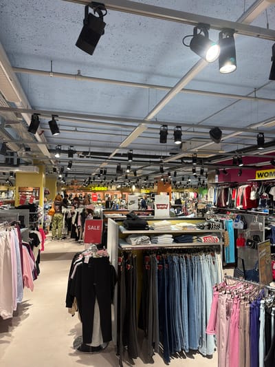 The image depicts the interior of a clothing store in Spreitenbach, Switzerland. A grid of metal beams and track lighting fixtures covers the ceiling. Rows of clothing racks display various items, including jeans, sweaters, and athletic wear. A "SALE" sign is visible among the clothing. Shoppers are present, although details are obscured.
