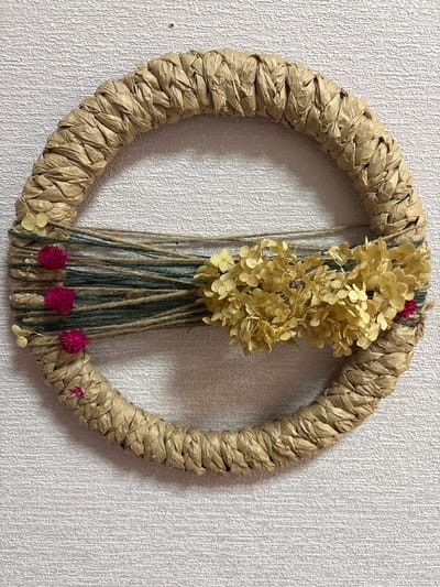This is a close-up, eye-level, medium shot of a circular wreath made of braided brown paper or straw, hanging on a white textured wall. The wreath is adorned with several bundles of dried yellow flowers and small pink flowers, tied together with twine and green string. The lighting is natural and even, casting subtle shadows on the wall. The overall impression is of a rustic, handmade decoration. The location is identified as Ветрынскі сельскі Савет, Belarus.