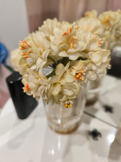 This media file features a close-up, slightly blurred shot of a bouquet of artificial light-yellow flowers with small orange accents in a clear glass vase. The flowers appear to be made of fabric and have delicate, layered petals. Some of the orange tips of the flower stamens are visible. The vase is filled with a light brown liquid.
The bouquet is placed on a white reflective surface, likely a table. To the left of the vase, a black, cylindrical object is partially visible; its function is unclear. In the background, a pinkish, sheer curtain is visible, suggesting an indoor setting. The lighting appears to be artificial, possibly from a room light, as there are some reflections on the surface. In the blurred background, there are hints of other objects, including another vase with flowers and what appear to be small, dark, blurry shapes on the reflective surface, possibly decorative spiders.
The overall mood of the image is quiet and still, with a focus on the floral arrangement. The shallow depth of field draws attention to the flowers, while the background elements are softly rendered. There is no visible text in the media file. The time of day and weather are not discernible. The location context of Staraya Kupavna, Russia, does not provide specific visual cues within the image itself.