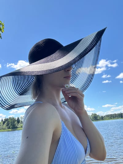 The image shows a medium shot of a woman wearing a large, black and white striped sun hat and a blue and white striped bikini top. She is holding a small white daisy up to her chin with her right hand. The woman is positioned in front of a body of water, likely a lake, with a treeline in the background and a blue sky with white clouds above. The location is identified as Lendži, Latvia. The overall scene suggests a bright, sunny day at a lakeside.