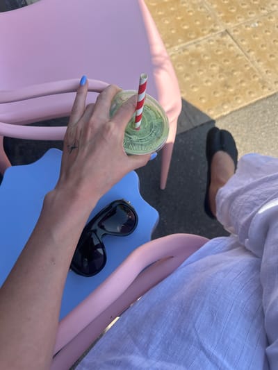 The image presents an overhead view of a person seated outdoors, possibly at a cafe or patio in Waverley, United Kingdom. The subject wears a light-colored, likely white, striped linen garment and black split-toe flat shoes. Their left hand, featuring light blue nail polish and a small, dark, abstract tattoo on the outer wrist, holds a clear plastic cup. The cup contains a green beverage and is fitted with a red and white striped straw. Text partially visible on the cup's lid includes "MP", "ABLE ON...", and "VEGETABLE", indicating the contents are likely a vegetable-based drink.
The person is seated on a colorful chair; a plain pink chair is visible on the left, while the person's right hand rests near black sunglasses placed on a wavy-edged blue surface, possibly a small table or part of another chair. The ground beneath consists of light tan tactile paving with raised bumps, next to darker gray paved sections, typical of public sidewalks or outdoor seating areas. Bright sunlight casts distinct shadows, suggesting a clear, sunny daytime. The overall scene depicts a relaxed moment, with the person enjoying a refreshment outdoors.