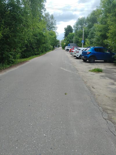 The image shows a street in Drezna, Russia, with parked cars along the right side. The street is paved and appears to be a two-lane road, though only the right lane and shoulder are visible. Dense green trees line the left side of the road, creating a canopy effect. On the right side, several cars are parked parallel to the road on a dirt and gravel shoulder. These vehicles include a blue hatchback, a white hatchback, a red hatchback, and a white SUV. A blue traffic sign with a white arrow pointing upwards is visible near the parked cars. The sky is overcast with clouds. Further down the road, more trees and buildings are visible in the distance. A person can be seen walking on the sidewalk in the distance on the left side of the road.