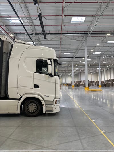 A white semi-truck cab is parked in a large, brightly lit warehouse in Sosnowiec, Poland. The truck is positioned in the foreground, taking up the left half of the image, with its side visible. The warehouse interior extends into the background, showing rows of shelves stacked with boxes. The floor is concrete, marked with yellow lines and numerous small white squares, some of which appear to be barcode labels. The ceiling is high, with exposed beams, fluorescent lighting, and skylights. There are no people visible in the image, and no apparent activity is taking place. The environment appears to be a logistics or distribution center.