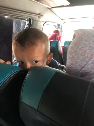 A close-up, slightly off-kilter shot captures a baby's face pressed against a black leather seat with teal trim. The baby has fair skin, dark hair, and dark eyes. The baby is looking directly at the camera. The background shows a window and other seats with a person wearing a pink shirt. The location is inside a vehicle, likely in Meunasahleubok, Indonesia. The focus is primarily on the baby's face.