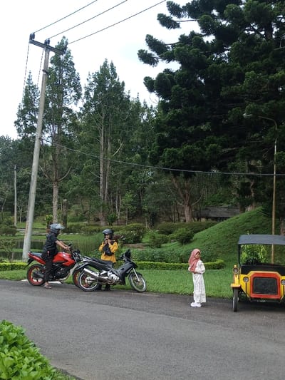The scene depicts an outdoor environment in Ambositra, Madagascar, featuring a paved road alongside a landscaped area. Three individuals and three vehicles are present. On the left, an individual wearing a dark shirt and a grey full-face helmet is seated stationary on a red sport motorcycle. Adjacent to this, a second individual in a yellow shirt and dark full-face helmet stands between the red motorcycle and a black scooter-style motorcycle, appearing to adjust something on their person. To the far right, a young girl in a light-colored dress and pink hijab stands facing left, positioned near a small, yellow open-sided recreational vehicle with an orange front. The background consists of a manicured grassy area with shrubs, leading to a dense forest of tall trees under an overcast sky. A utility pole with multiple power lines is visible on the left, and a pond or water feature is discernible behind the motorcycles.
