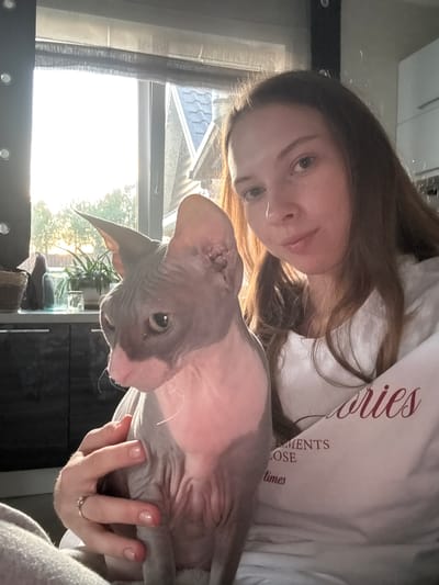 A young woman with long brown hair is holding a hairless cat in front of a window with bright sunlight streaming through. The woman is wearing a white t-shirt with text that reads "MEMORIES LOSE times". The cat is grey and pink and looking to the left. In the background, there is a kitchen counter with plants and jars, and a glimpse of a roof and trees outside the window. The scene appears to be indoors, likely in a residential setting in Novaya Ivanovka, Russia.