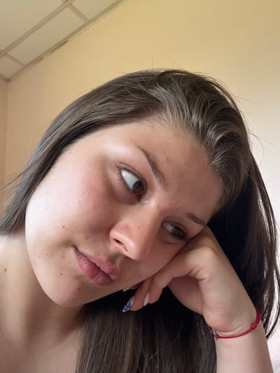 A close-up, low-angle selfie of a young woman with brown hair, resting her head on her hand. Her right hand, adorned with a red string bracelet and patterned nail polish, supports her cheek. Her gaze is directed off to the side, away from the camera. The background consists of a plain, light-colored wall and a ceiling with acoustic tiles. The scene appears to be indoors. The location in the city of Prodanovtsi, Bulgaria is not discernible from the image itself.