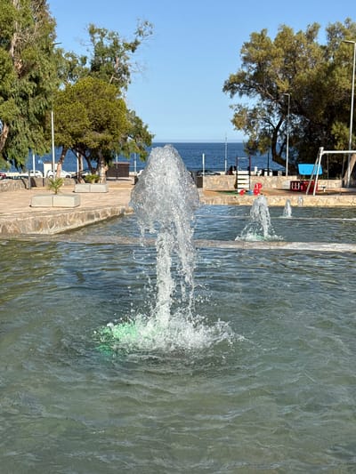 The image shows a water fountain with several jets spraying water. The fountain is set in an outdoor area with a paved walkway and stone borders. In the background, there are trees, the sea, and a clear blue sky. Some lounge chairs and tables are visible near the sea. The time of day appears to be daytime, with bright sunlight casting shadows. The weather seems pleasant and sunny. The scene suggests a resort or public area near the coast in Aguadulce, Spain, likely a place for relaxation and leisure. There is no visible text in the image.