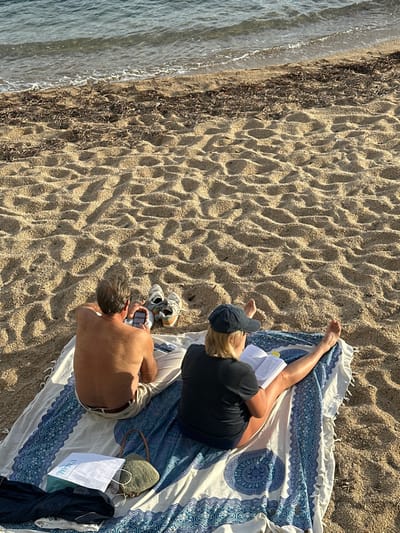 On a sandy beach in Palafrugell, Spain, two individuals are seated on a patterned blue and white blanket, observed from behind. The male, positioned on the left, is shirtless and wears light-colored trousers, holding and interacting with a smartphone. Behind him on the sand is a pair of light-colored sneakers. The female, on the right, wears a dark t-shirt and a dark baseball cap, reading an open book. Her bare legs are extended on the blanket. Adjacent to the male's left side on the blanket are a greenish-beige woven bag, a white paper bag, and a dark piece of fabric. The sandy environment features ripples and depressions, with a line of dark organic debris along the waterline, beyond which calm, light blue-green ocean water is visible under a warm, low-angle light.