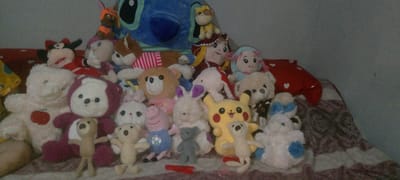 The image showcases a collection of stuffed animals arranged on a patterned blanket. The stuffed animals include a large blue Stitch, several teddy bears of various sizes and colors, a Mickey Mouse, a pig wearing a soccer jersey, a yellow Pikachu, and other animal characters with ears. Some of the stuffed animals are arranged in front of others, creating a layered composition. A red pillow with white hearts is visible to the right of the collection. The background is a plain, light-colored wall. The lighting appears to be artificial, suggesting the photograph was taken indoors. There is no discernible activity or event taking place, as the stuffed animals are static. The overall impression is a display of toys, possibly in a child's room or a collector's space. There is some text visible on a white teddy bear which says "good" and on the pig toy which is difficult to read, it is a soccer ball with colors. There are no people visible in the image. The location context of Marhamat, Uzbekistan is not visually apparent from the image itself.