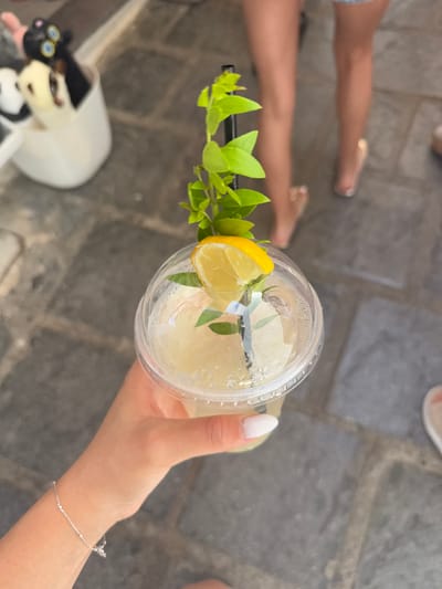 In Lindos, Greece, a person's hand, adorned with a delicate silver bracelet and a white-tipped manicure, holds a clear plastic cup filled with a pale yellow beverage. The drink is garnished with a slice of lemon, sprigs of fresh green leaves, and a black straw. The cup is sealed with a clear plastic lid, and condensation has formed on its surface. The background is a textured gray stone pathway, with blurred figures of people in the distance, suggesting an outdoor setting. To the left, a white container holds several plush toys, including a black one with large blue eyes and a cream-colored one. The scene captures a moment of refreshment, likely in a tourist area.