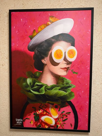 The image shows a framed, surrealist painting of a woman. Her eyes are covered by two halves of a hard-boiled egg, showing the yolk and white. She wears a large, ruffled collar made of green lettuce leaves around her neck. On her head, she has a white hat adorned with what appears to be a croissant and other food items. A sprig of green herb is held delicately in her mouth. The background is a vibrant, textured pink. Below the woman's neck, the painting depicts a bowl of soup or salad, also with visible egg pieces and herbs. In the bottom left corner of the painting, there is a signature in white cursive script, which reads "Šaltibarščių portretas" (Cold beet soup portrait). Beneath this, there is another line of text, possibly an artist's name and date, that appears to be "Andrius Dzinga (20/30)" or similar. The painting is hanging on a textured, light-colored wall. The text in the bottom left corner of the painting indicates that this artwork is located in Rūdiškės, Lithuania.