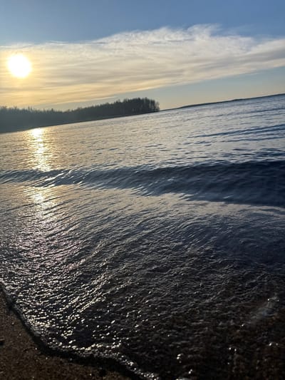 The image shows a sunset over a body of water, likely a lake, with the sun low on the horizon. The shoreline is visible on the right edge of the frame, where the water meets the sand. The water's surface is rippled with waves, reflecting the sunlight. In the distance, a dark line of trees and land are visible across the water. The sky is a mix of blue and white clouds, with a golden hue from the setting sun.
It is not possible to identify the exact location in the city of Glubokoe, Russia based on this image alone.