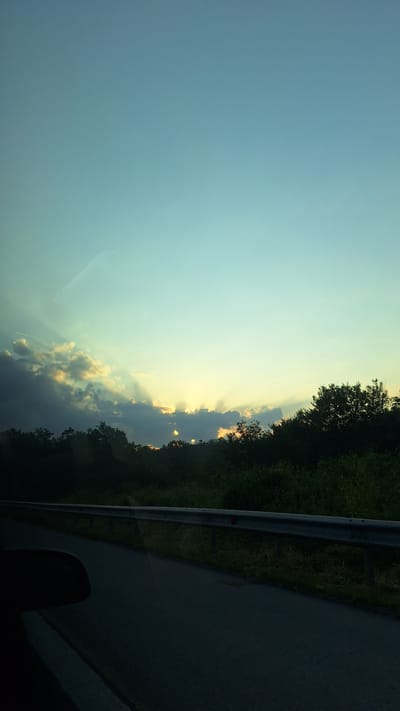 The image captures a scene from a moving vehicle, likely on a road in or near Ravnishte, Bulgaria, given the context provided. The perspective is from the passenger seat, looking out the window. The time of day appears to be late afternoon or early evening, as evidenced by the dramatic crepuscular rays of sunlight streaming through the clouds.
The sky dominates the upper portion of the frame, transitioning from a light, clear cyan at the top to a softer, warmer hue closer to the horizon. A few wispy clouds are visible, with darker, more substantial clouds on the left side where the sun's rays are most prominent. These rays are sharply defined, suggesting they are passing through gaps in the cloud cover.
Below the sky, a dense line of dark green trees forms the horizon, suggesting a rural or semi-rural setting. The trees appear to be a mix of deciduous and possibly some evergreen varieties, forming a thick canopy.
In the foreground, a dark asphalt road curves gently to the right, indicating the vehicle's path. A metal guardrail runs along the side of the road, separating it from the grassy verge and the trees beyond. The guardrail appears to be made of silver-colored metal posts and rails, with some subtle signs of wear and possibly some grass growing around its base.
On the left side of the frame, a dark, shadowy object is visible in the lower corner. This is likely the interior of the vehicle, possibly a car door or a side mirror, captured in silhouette. There is also a faint reflection on the window, a common occurrence when shooting from inside a vehicle. No people are visible in the image, and no specific actions or interactions are depicted beyond the implied movement of the vehicle. The overall environment suggests a roadside view during a scenic sunset.