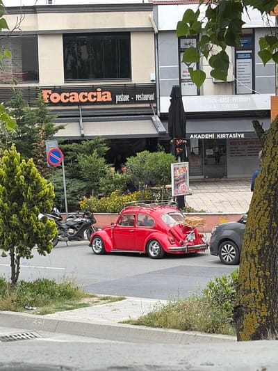 The image shows a street in Esenler, Turkey. On the left is a building with a restaurant called "Focaccia pastane restaurant" and a business providing "TİCARET SICIL DANIŞMANLIĞI" services. Next to it is "AKADEMİ KIRTASİYE" selling stationery. A red vintage car is parked on the street, next to a parked black motorcycle. There's a blue and red parking sign nearby. Pedestrians are visible in the background. A tree trunk and branches partially obscure the view on the right and left of the image.