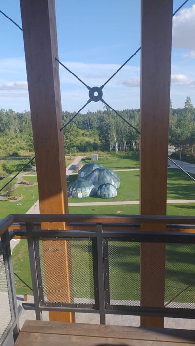 The image shows an aerial view of the Tērvete Nature Park in Tērvete, Latvia, taken from a wooden observation tower. In the center of the frame are several large, green, inflatable dome-like structures, resembling bubbles, scattered across a grassy field. The domes have what appear to be images of butterflies on some of them. A paved pathway winds through the park, and benches are visible. In the background, a dense forest of deciduous and coniferous trees stretches towards the horizon under a bright blue sky with scattered clouds. The foreground of the image features the wooden beams and railings of the observation tower, with wooden planks visible at the very bottom of the frame.
