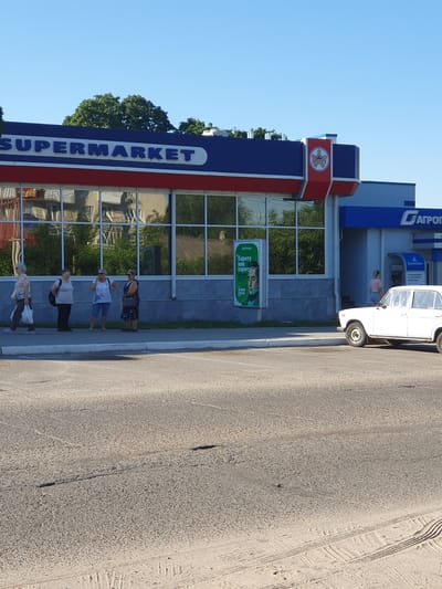 In Dubossary, Moldova, a supermarket named "SHERIFF" with a prominent red and blue facade stands under a clear blue sky. Four women are gathered on the sidewalk in front of the building, engaged in conversation. To the right of the supermarket, a white car is parked, and a woman is seen entering a building with an ATM. A billboard advertising "Sberbank" is displayed on the exterior wall. The scene unfolds on a sunny day, with trees visible in the background, reflecting in the supermarket's windows. The foreground consists of a paved road.
