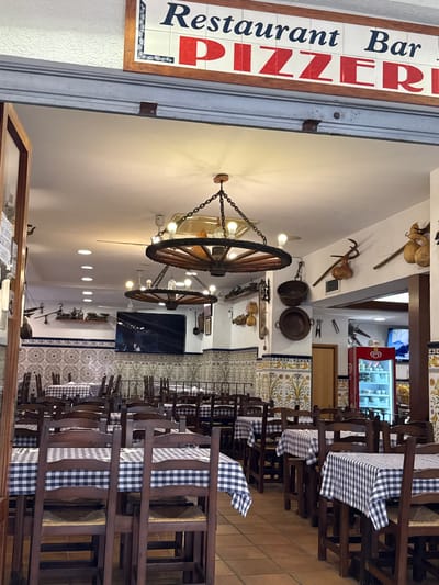 The image shows the interior of a restaurant and bar, identified by a sign above the entrance that reads "Restaurant Bar PIZZERIA". The restaurant is decorated with traditional tiles on the walls and features wooden tables covered with blue and white checkered tablecloths. The furniture appears to be made of dark wood. Several light fixtures are visible, including a large, rustic chandelier that resembles a wagon wheel with exposed bulbs. Decorative items such as old farming tools, pots, and gourds are displayed on the walls. A large television screen is mounted on the wall, displaying an image. In the background, a red refrigerator with a Coca-Cola logo is visible. The restaurant is empty of people, suggesting it is either closed or the photo was taken before opening hours. The overall impression is a cozy, rustic, and traditional dining establishment.