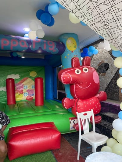 The image shows a Peppa Pig-themed inflatable bouncy house set up indoors at a venue in Bonadikombo, Cameroon (latitude: 4.048, longitude: 9.218). The bouncy house is partially inflated, revealing its internal structure and the cartoon imagery printed on its walls. Two red inflatable columns stand on the green play area. A large, red inflatable figure of Peppa Pig, in her signature polka-dot dress, is standing near the bouncy house. A white plastic chair sits next to the bouncy house. In the background, there are white, blue, and yellow balloons hanging from the ceiling, which is decorated with a black and white grid pattern. A person, partially visible in the bottom left of the image, is standing in front of the bouncy house.