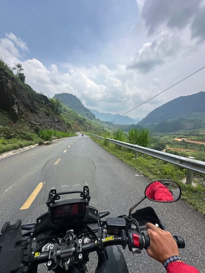 The image presents a first-person perspective from a motorcycle rider on a mountain road. The rider's right hand, wearing a bracelet with a blue and white pattern, grips the throttle, with a rear-view mirror reflecting a portion of the rider's red jacket. The black handlebars, windshield, and dashboard of the motorcycle occupy the lower right foreground.
The asphalt road stretches ahead, marked with a continuous yellow line on the left and broken yellow lines towards the center, leading into a distant curve. A metal guardrail runs along the right side of the road, bordered by lush green grass and dense vegetation on a steep, rocky mountainside. In the midground and background, multiple layers of green, forested mountains rise under a bright, partly cloudy sky, with some power lines visible overhead. The scene suggests a journey through a rugged, natural landscape.
The setting is a winding mountain pass, consistent with the provided context of Minh Sơn Commune, Vietnam. It is daytime, with bright natural lighting. Visible text on the motorcycle includes "SPIRIT BEAST" on the handlebar clamps, and "FRON AS ONLY DOT BRAKE FLUID", "FILLER CAD CONTAINER CLEAN", and "FREIN DOUR" on a brake fluid reservoir or related component.