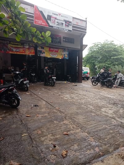 The scene is an outdoor, paved area in front of a building. The building is a motorcycle shop called "Banca Buaya Motor" and is located in Kecamatan Caringin, Indonesia, at latitude -7.523 and longitude 107.485. The shop is selling Yuasa batteries and has signage for "Aspira" and "Astra Quality". The scene appears to be a parking lot, with several motorcycles parked in front of the shop. Some people are gathered near the motorcycles, while others are entering the shop. The weather is overcast and there are some leaves on the ground.  A white van is parked behind the motorcycles.