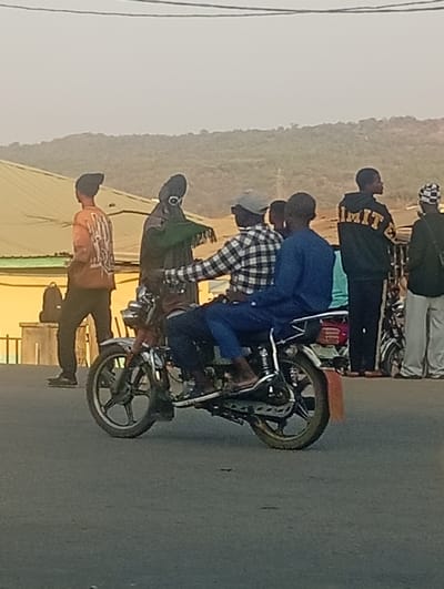 A street-level photograph captures a moment of daily life at Itakpe Junction, Nigeria, under a hazy sky. The central focus is a red and silver motorcycle on a paved road, occupied by two men. The driver, wearing a plaid shirt and a grey cap, is positioned forward, while his passenger, dressed in blue, sits behind him.

Several other individuals populate the scene. To the left, a person in an orange long-sleeved shirt and a dark beanie walks away from the viewer, carrying a dark bag over their shoulder. Behind the motorcycle, another figure in a dark green garment and an unusual, possibly hooded, head covering stands. On the right side of the frame, a man in a black hoodie with "LIMIT" in yellow text across the back stands, looking towards the right. Further right, a person in white pants and a patterned head covering is partially visible.

The background reveals parts of buildings with light-colored roofs, suggesting an urban or semi-urban setting, and distant hills covered in greenery, indicating a hilly terrain. Overhead utility wires traverse the upper portion of the image. The lighting is soft and ambient, characteristic of either early morning or late afternoon, with no harsh shadows. The overall impression is one of casual activity at a busy junction, with people either moving or gathered, reflecting the everyday rhythm of the location.