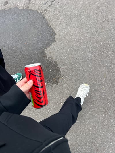 A first-person perspective photograph shows a person holding a red Coca-Cola can. The person is wearing black pants and a black jacket. The person is also wearing one white sneaker and one green sneaker with white stripes. The person is standing on a gray asphalt surface. The person took this photo in Noisiel, France.