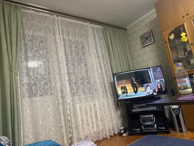 A photograph taken indoors, seemingly in a residential setting in Vorotynsk, Russia. The dominant features include a television playing what appears to be a stage performance, flanked by long curtains that are partially drawn. To the right of the television, a portion of a cabinet is visible, as well as a table holding a bottle of milk labeled "Capapaново" (Sarapanovo) and other unseen items. Below the television is a multi-tiered entertainment center containing various electronic devices, including a stereo system. A portion of bedding is visible in the foreground, suggesting the image was taken from a bedroom or living room. The wallpaper is patterned. A photograph of a group of people, possibly family members, hangs on the wall.